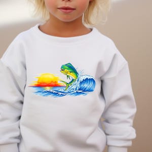 May include: White sweatshirt featuring a colorful graphic of a mahi-mahi fish leaping from a wave in front of a setting sun. The fish is yellow and green, the waves are blue, and the sun is yellow and orange. A children's apparel item.