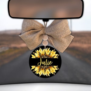 May include: A round car air freshener with a black background and a yellow sunflower with a leopard print center. The name "Julie" is written in yellow across the center of the sunflower.