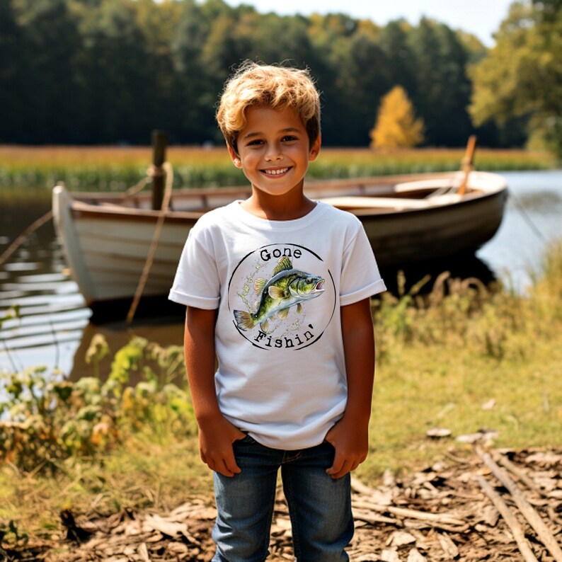 May include: White t-shirt featuring a fish graphic and the text "Gone Fishin'". A child in blue jeans smiles in front of a lake with a boat. The image captures a sunny day and the theme of fishing.
