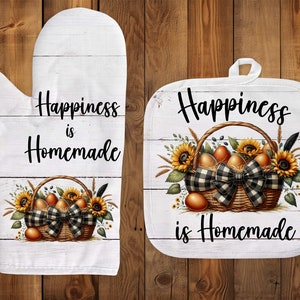 Puede incluir: Un guante de horno blanco y un posavasos blanco con fondo de madera. Ambos tienen el texto "Happiness is Homemade" y una cesta de girasoles y huevos.