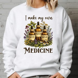 May include: White sweatshirt with the text "I make my own MEDICINE." The design features three amber tincture bottles surrounded by colorful flowers and herbs. The bottles are labeled "Goldenseal Tincture," "Valerian Root Tincture," and "Turkey Tail Tincture."