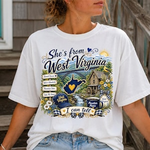 May include: A white t-shirt featuring a graphic print with the text She's from West Virginia I can tell. The design includes a bridge, a water mill, a map of West Virginia with a yellow heart, a blue baseball cap, and a mug labeled Mountain Mama.