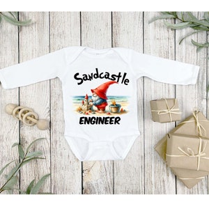 May include: White long-sleeved baby onesie with a graphic of a gnome building a sandcastle on a beach. The text "Sandcastle Engineer" is printed below the graphic.