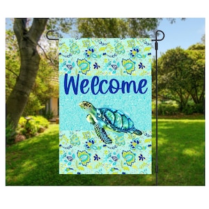 May include: A garden flag featuring a watercolor sea turtle design. The flag has a turquoise background with the word "Welcome" in blue. The flag is decorated with a floral pattern in blue and yellow.