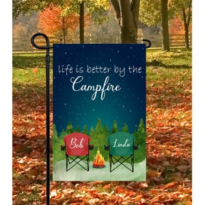 Puede incluir: Una bandera de jardín con un fondo de cielo nocturno azul estrellado. El texto "Life is better by the campfire" está escrito en blanco. Dos sillas de camping están frente a una fogata con los nombres "Bob" y "Linda" escritos en ellas.