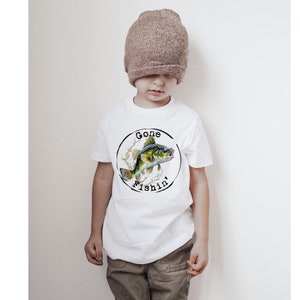 May include: White t-shirt with a circular graphic showing a fish illustration and the words "Gone Fishin'". The fish is green, yellow, and blue, with water splashes. A child is wearing the shirt and a brown beanie.