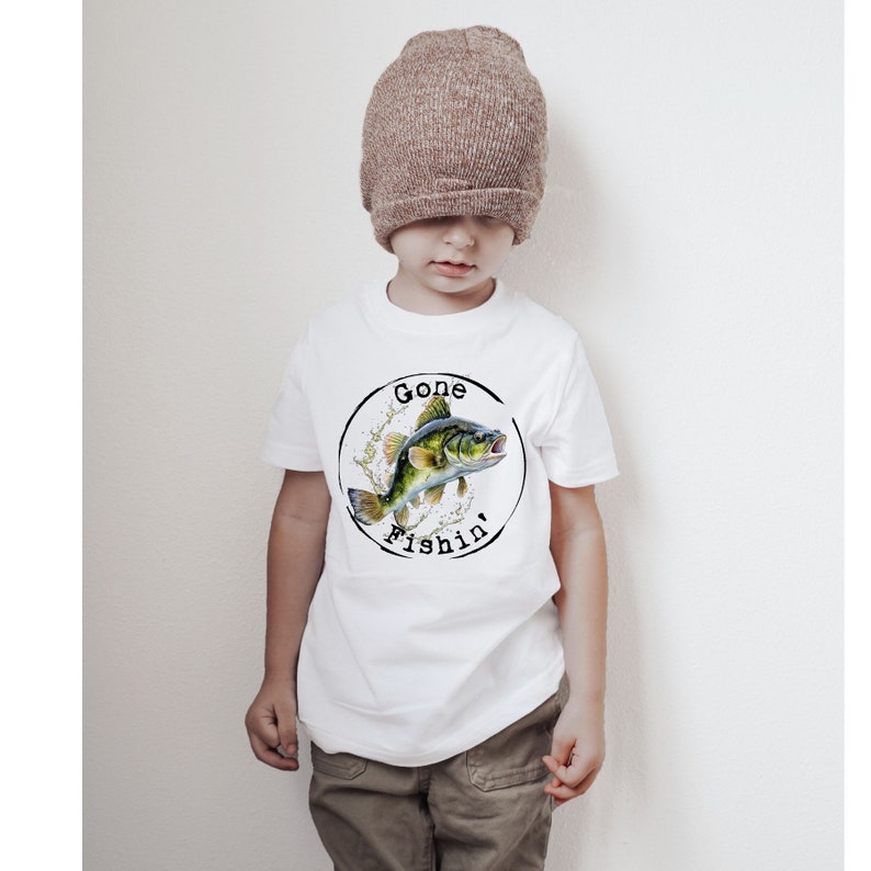 May include: White t-shirt with a circular graphic showing a fish illustration and the words "Gone Fishin'". The fish is green, yellow, and blue, with water splashes. A child is wearing the shirt and a brown beanie.