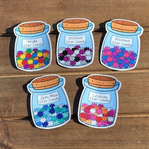 May include: Five blue glass jars with cork lids filled with colorful star-shaped candies. Each jar has a label with a different identity label: Pride, Asexual, Bisexual, Gay Men, and Lesbian. The jars are arranged in a row on a wooden surface.