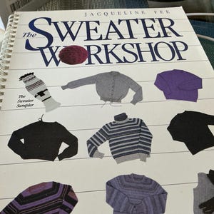 May include: A spiral-bound book titled "The Sweater Workshop" by Jacqueline Fee. The cover features illustrations of various sweaters in different colors and styles, including a gray cardigan, a purple crewneck, and a striped turtleneck. A ball of yarn is also depicted.
