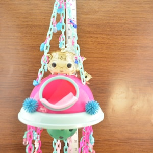 May include: A pink and blue bird toy with a gold birdhouse and a variety of colorful chains and beads. The toy is hanging from the ceiling.