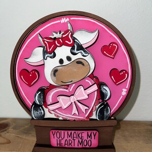 May include: A wooden Valentine's Day decoration featuring a cow holding a heart-shaped gift box. The cow has black and white spots, a red bow, and is set against a pink background with red hearts. The base reads "YOU MAKE MY HEART MOO."