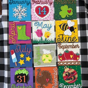 May include: Twelve painted wooden squares, each representing a month of the year. Each square features a unique design and color scheme, such as a snowflake for January, a heart for February, a shamrock for March, and a turkey for November.