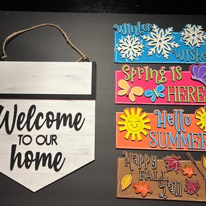 May include: A set of five wooden signs with interchangeable seasonal greetings. The signs feature a variety of colors and designs, including snowflakes, butterflies, suns, and leaves. The signs read "Winter Wishes", "Spring is Here", "Hello Summer", and "Happy Fall Y'all".