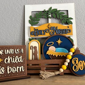 May include: Wooden Christmas decorations with a nativity scene, a star, and the words "For unto us a child is born" and "Holy Night". The decorations are painted in shades of blue, yellow, and brown.