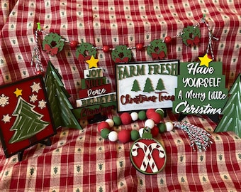 Könnte beinhalten: Eine Sammlung von weihnachtlichen Holzschildern mit verschiedenen Sprüchen, darunter "Farm Fresh Christmas Trees", "Have Yourself A Merry Little Christmas" und "Joy Noel Peace Believe Christmas". Die Schilder sind in Grün und Rot dekoriert und zeigen einen Stern, Schneeflocken und einen Zuckerstangen-Anhänger.