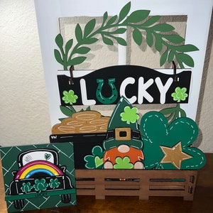 May include: A St. Patrick's Day themed wooden display featuring a "Lucky" sign, a leprechaun, a pot of gold, and a shamrock. A small square sign shows a truck with a rainbow and the word "Lucky". The display is on a wooden surface.