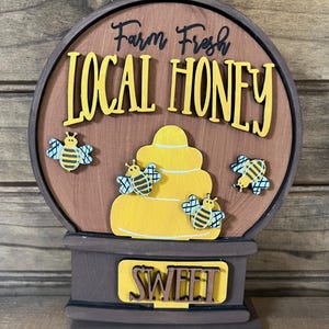 May include: A wooden sign shaped like a honeybee hive with the text "Farm Fresh Local Honey" and "Sweet" on it. The sign is decorated with four yellow and black honeybees.