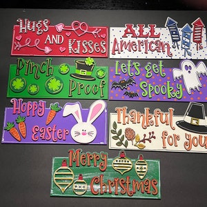 May include: A set of eight wooden signs with various holiday themes. The signs feature painted designs and text, including "Hugs and Kisses", "All American", "Pinch Proof", "Let's get Spooky", "Hoppy Easter", "Thankful for you", and "Merry Christmas".