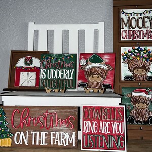 May include: A set of six wooden signs with Christmas themes featuring a red barn, a Highland cow, a Christmas tree, and the phrases "Christmas is Udderly Delightful", "Mooey Christmas", "Christmas on the Farm", and "Cowbells Ring Are You Listening".