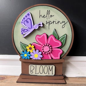 May include: A wooden spring-themed decoration featuring a purple butterfly and pink flower. The words "hello spring" are written on the green background. The word "BLOOM" is on the base. The decoration is brown and has a round shape.