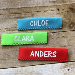 May include: Three rectangular, colorful fabric items with white text spelling out names. The top one is teal and says "CHLOE", the middle one is lime green and says "CLARA", and the bottom one is red and says "ANDERS".