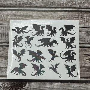 May include: A white rectangular sheet featuring a collection of fifteen individual black dragon silhouettes in various poses, including flying, standing, and crouching positions, arranged in a grid pattern across the surface.
