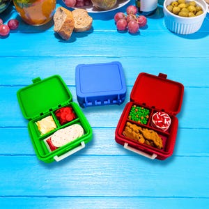 May include: Three square plastic lunch containers in bright green, blue, and red. The green container is open, holding crackers, cheese, and red slices. The red container is open, holding green peas, a round treat, and brown snacks.