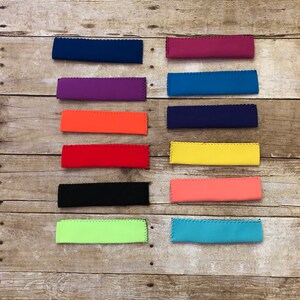 May include: A collection of colorful, rectangular neoprene drink sleeves in various shades, including blue, purple, orange, red, black, green, yellow, and pink. Each sleeve has a scalloped edge and is designed to insulate beverages.