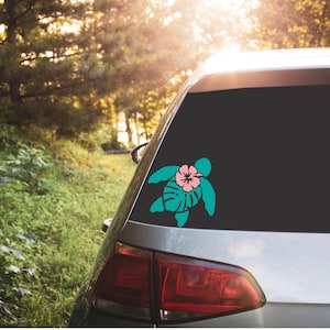 May include: A teal turtle decal with a pink hibiscus flower and green leaf accents is affixed to the rear window of a silver car. The car is parked outdoors, with a backdrop of lush green foliage and sunlight.