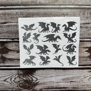May include: A white rectangular sheet featuring sixteen individual black dragon silhouettes in various poses, including flying, standing, and crouching positions, arranged in a grid pattern on a clear adhesive backing.