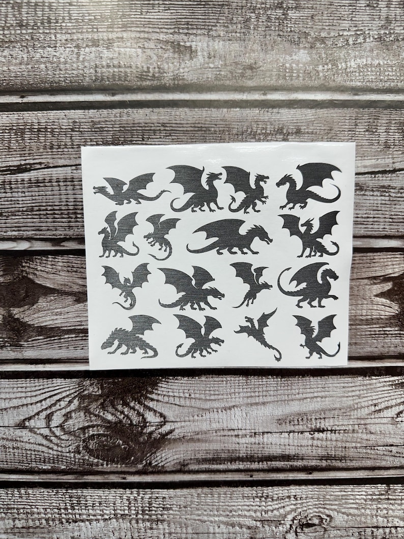 May include: A white rectangular sheet featuring sixteen individual black dragon silhouettes in various poses, including flying, standing, and crouching positions, arranged in a grid pattern on a clear adhesive backing.