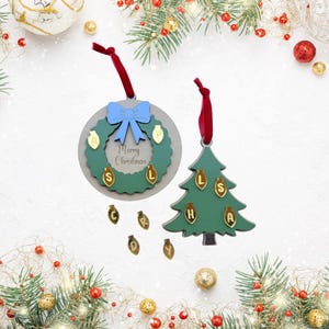 May include: Two Christmas ornaments with red ribbons: a circular grey plaque featuring a green wreath, a blue bow, and the text Merry Christmas, and a green tree-shaped ornament. Both feature small gold light bulb charms with letters.