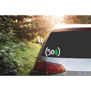 May include: A white vinyl decal applied to a car rear window featuring the number 50 in white, followed by a green pine tree icon, all enclosed within two white parentheses. The design is centered on the dark glass of the vehicle window.