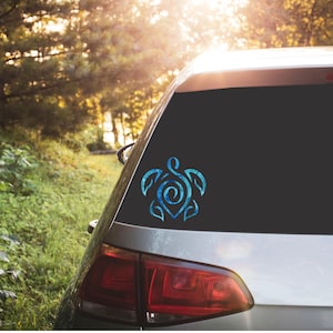 May include: A blue sea turtle decal with a spiral shell pattern applied to the rear window of a silver car. The turtle design features a stylized, abstract shape with light and dark blue tones, creating a marbled or water-like appearance.