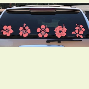 May include: A set of five coral-coloured hibiscus flower decals applied to the rear window of a vehicle. Each flower features a distinct floral design with visible petals, a central stamen, and leaves, arranged horizontally across the glass.