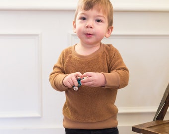 Puede incluir: Un niño pequeño con un suéter marrón y pantalones vaqueros azules está de pie frente a una pared blanca. El niño sostiene un pequeño juguete en sus manos.