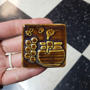 May include: A square, brown ceramic tile with a honey theme. The tile features embossed images of honeycombs, a honey pot with dripping honey, and two bees. The tile is held in a hand.