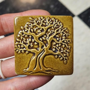 May include: A square, ceramic tile with a raised relief of a tree design. The tree has a thick trunk and branches with detailed leaves. The tile is a warm, earthy tone with a glossy finish.
