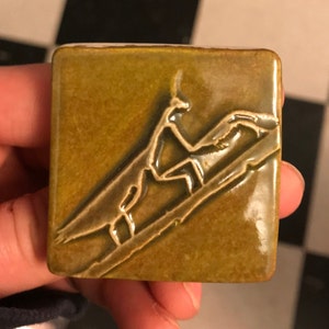 May include: A square ceramic tile with a green glaze. The tile features a raised design of three stylized birds in flight.