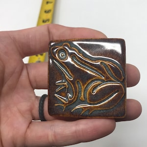 May include: A square, ceramic tile with a raised relief frog design. The tile is a dark brown color with a glossy finish. The frog is a lighter brown color with gray accents. The tile is held in a hand.
