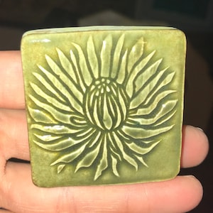 May include: A green ceramic tile with a raised design of a stylized flower. The flower has a central bud and many petals radiating outwards.