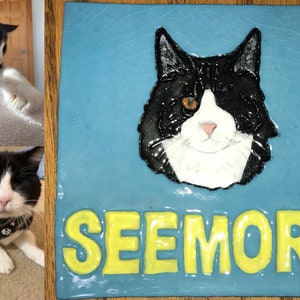 May include: A blue ceramic tile with a painted portrait of a black and white cat with one orange eye. The cat's name, "SEEMORE", is written in yellow letters below the portrait.