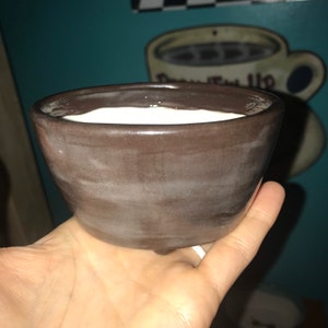 May include: A small, brown ceramic bowl with a white interior. The bowl is slightly tilted and has a subtle, uneven glaze.
