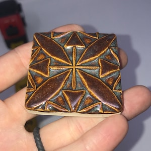 May include: A square ceramic tile with a geometric pattern in shades of brown and blue. The tile has a raised design and a glossy finish.