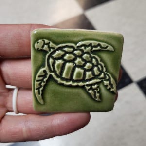 May include: A square, green ceramic tile featuring a raised design of a sea turtle. The turtle is detailed with dark green accents, and the tile has a glossy finish. The tile is approximately 2 inches square.