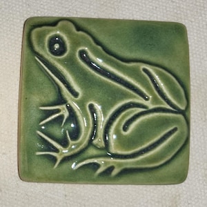 May include: A green ceramic tile with a raised frog design. The frog is outlined in black and has a glossy finish.