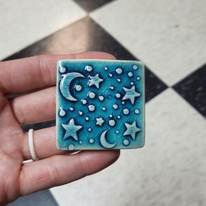 May include: A square, turquoise ceramic tile featuring a celestial design. The tile has a crackle glaze and is decorated with white crescent moons, stars, and small dots. The tile is held in a hand.