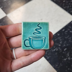 May include: A square, teal ceramic tile with a coffee cup design. The cup is light blue with steam rising from it. The tile has a crackled texture and is held in a hand.