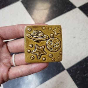 May include: A square, ceramic tile with a space theme. The tile is a mustard yellow color and features a planet with rings, a crescent moon, two stars, and small circular accents. The design is raised and has a textured appearance.