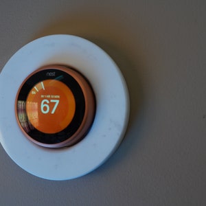May include: A smart thermostat with a digital display showing the current temperature of 67 degrees Fahrenheit. The thermostat is round with a copper-colored ring and is mounted on a white circular base. The display shows the temperature in an orange color.